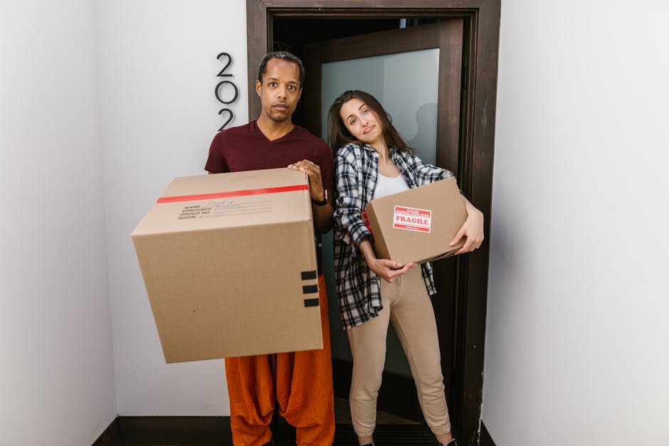 A man with dark skin, curly black hair, and a beard is inside a bright, minimally furnished room with white walls and wooden flooring, engaged in a house removal process. He is wearing a blue T-shirt 