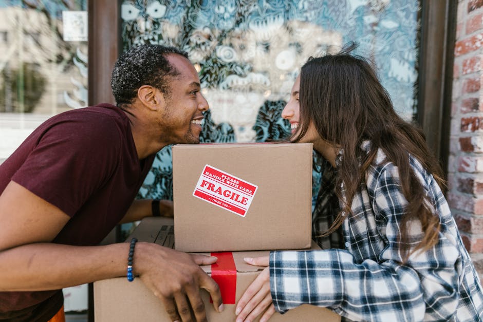 A person wearing light-colored clothing and white socks is in the process of packing or preparing for a home relocation, placing a cardboard box on top of other sealed boxes inside a room with wooden