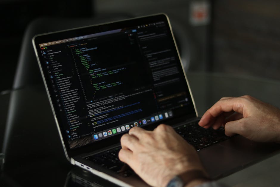 A close-up of a person's right hand typing on a silver laptop keyboard, placed on a wooden surface. The person is wearing a wristwatch with a black strap on their left wrist, and the laptop screen dis